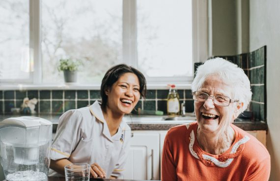 An elderly woman laughs beside a friendly young care assistant A young, friendly and bubbly carer sits beside an elderly woman at a kitchen table. The older woman faces the camera directly and laughs as the younger woman laughs behind her. The scene is lighthearted and fun, and the older woman quite clearly has a big personality.